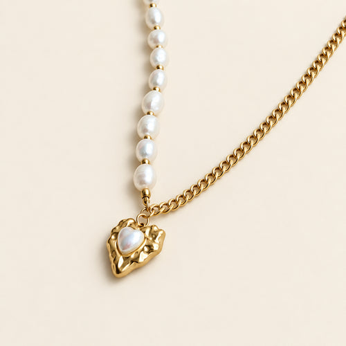A gold chain necklace featuring a row of white baroque pearls leading to a textured gold heart pendant with a white inlay, displayed on a soft beige surface with white feathers.